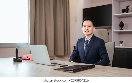 Portrait Of Happy Asian Businessman CEO In Suit With Sitting At Work Desk And Looking At Camera In Modern Room At Workplace. Handsome Male Executive Financial Director Smile And Successful In Office.