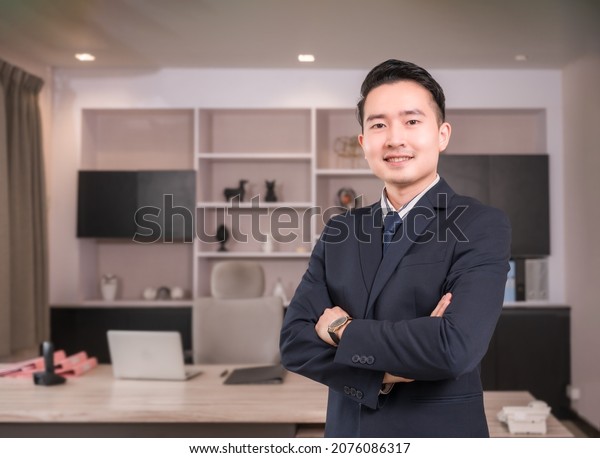 Portrait of happy Asian businessman CEO in suit\
with arms crossed and looking at camera in modern room at\
workplace. Handsome male executive financial director standing and\
smiling success in\
office.