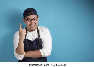 Portrait Of Happy Asian Chef Or Waiter Smiling With Pointing Finger Up, Having Bright Good Idea Gesture, Against Blue Background