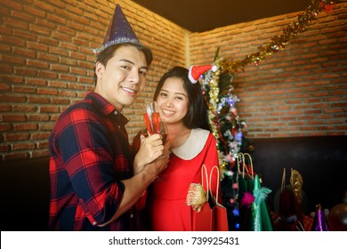 Portrait Of Happy Asian Couple, 20 To 30 Years Old, With Santa Hats Holding Hands And Toasting Glasses Of Red Wine To Celebrate Christmas Or Xmas And New Year Party.