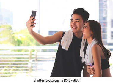 Portrait Of Happy Asian Couple Wearing Sportswear With Towel Around Neck, Relax After Workout, Standing Outdoor And Taking Selfie Photos With Smartphone. Sport And Health Concept.