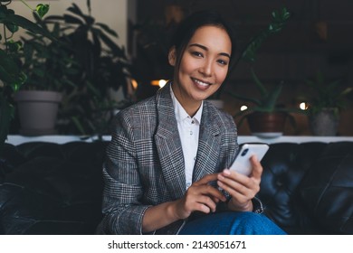 Portrait of happy Asian female blogger with digital mobile device smiling at camera during leisure pastime, successful business woman in smart casual apparel holding smartphone gadget and posing