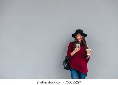Portrait of a happy asian girl dressed in hat and sweater holding coffee cup while standing and using mobile phone on a city street
