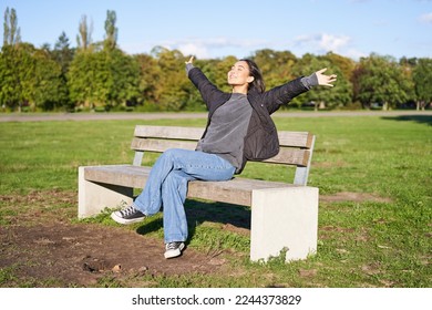 Portrait of happy asian girl feeling freedom and excitement, stretching hands while sitting on bench and smiling.