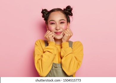 Portrait of happy Asian girl with pinup makeup, dark hair combed in two buns, piercing in nose, wears casual yellow sweatshirt and overalls, stands on pink background, feels happy. Ethnicity, emotions - Powered by Shutterstock