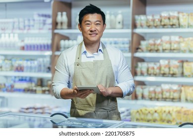 Portrait Of Happy Asian Male Small Business Owner Standing In Grocery Store Behind Counter. Male Manager With A Tablet Looking At The Camera. Friendly Administrator Food Market. Worker In Apron