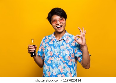 Portrait Of A Happy Asian Man Isolated Over Yellow Background, Holding Bottle With Fizzy Drink