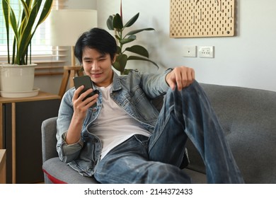 A Portrait Of A Happy Asian Man Lying Down On A Couch In A Living Room Using A Smart Phone, For Home And Technology Concept.