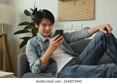 A Portrait Of A Happy Asian Man Lying Down On A Couch In A Living Room Using A Smart Phone, For Home And Technology Concept.