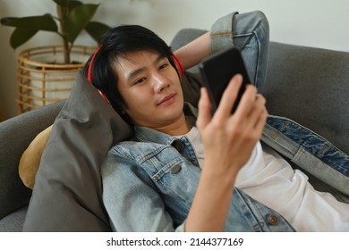 A Portrait Of A Happy Asian Man Lying Down On A Couch In A Living Room Using A Smart Phone For Entertainment, For Home And Technology Concept.