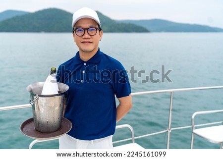Portrait of Happy Asian man waiter holding champagne in ice bucket for serving to passenger tourist travel on luxury catamaran boat yacht on summer vacation. Cruise ship service occupation concept.