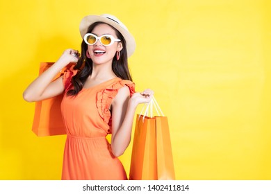 Portrait of a happy Asian pretty girl holding shopping orange bags away isolated over yellow background,colorful shopping concept.
