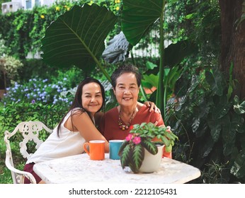 Portrait Of Happy Asian Senior Woman And Her Daughter  Sitting Together At White Table In Beautiful Garden, Daughter Hugging Her Mother Smiling And Looking At Camera.