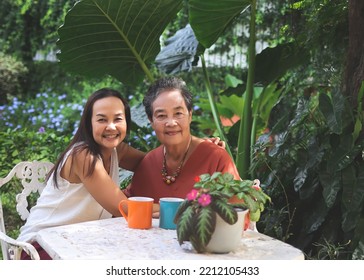 Portrait Of Happy Asian Senior Woman And Her Daughter  Sitting Together At White Table In Beautiful Garden, Daughter Hugging Her Mother Smiling And Looking At Camera.