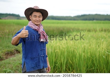 Portrait of happy Asian Thai male farmer wears cowboy hat and shows thumb up for good product at his green organic rice paddy field. Concept agricultural occupation. Satisfied in agricultural product.