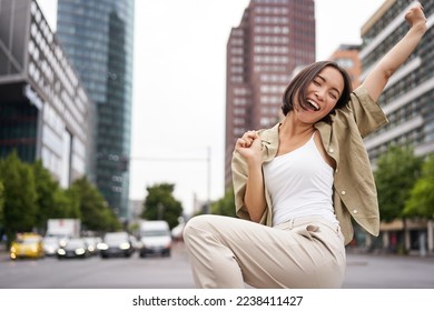 Portrait of happy asian woman, dancing and feeling joy, triumphing, raising hand up in victory gesture, celebrating on streets. - Powered by Shutterstock