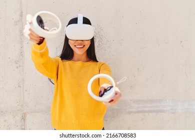 Portrait of happy asian woman gesturing with virtual reality hedset and controllers standing against concrete wall - Powered by Shutterstock