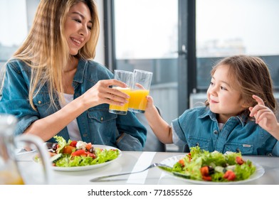 Portrait of happy asian woman and girl clinking glasses of juice and laughing. They are sitting at table and eating salad in kitchen Foto Stok