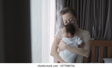 portrait of happy asian woman mother holding sleepy baby infant near window at home daytime. 4 months baby embracing in parent arm. relationship mother and daughter. mom touching baby lull to sleep.