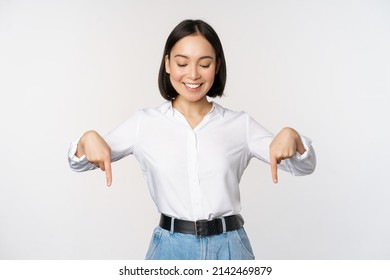 Portrait of happy asian woman pointing fingers down and looking below at advertisement, showing info banner, advertising, standing over white background - Shutterstock ID 2142469879