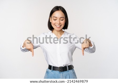 Portrait of happy asian woman pointing fingers down and looking below at advertisement, showing info banner, advertising, standing over white background