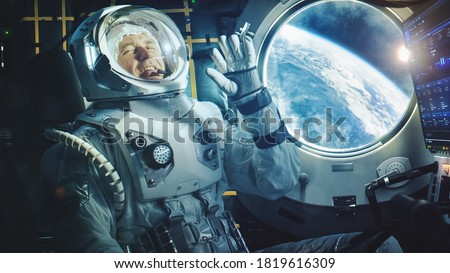 Portrait of a Happy Astronaut on a Space Ship In Orbit. Cosmonaut in a Futuristic Space Suit is Full of Joy and is Waving Hand on a Video Call. VFX Graphics Shot from the International Space Station.