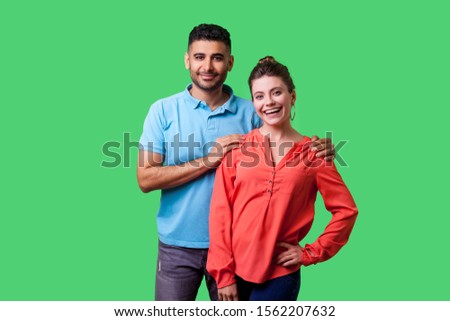 Portrait of happy attractive young couple in casual wear standing together, looking at camera with toothy smile, caring man holding female shoulders. isolated on green background, indoor studio shot
