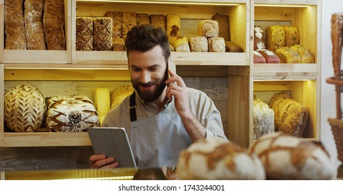 Portrait of happy baker working in bakery shop using digital tablet computer and talking on cell phone - Powered by Shutterstock