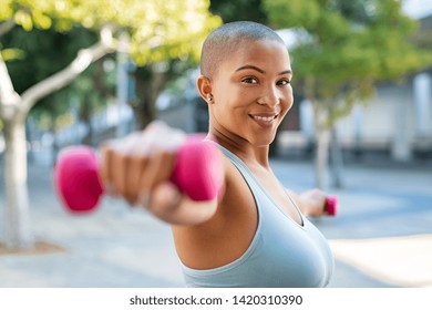 Portrait of happy bald woman exercising while looking at camera. Smiling girl doing sports outdoors with dumbbells lifting weights. Fit fitness girl in sportswear exercising outside to slim down.