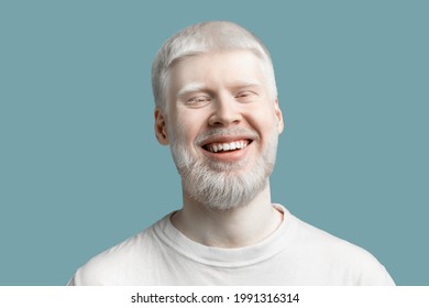 Portrait of happy bearded albino man with pale skin and white hair wearing t-shirt, posing and smiling to camera on turquoise studio background. Abnormal deviations, unusual appearance - Powered by Shutterstock