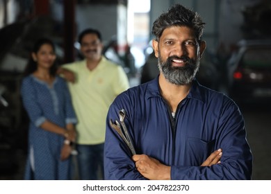 Portrait Of A Happy Bearded Car Mechanic Smiling In Car Service Station. Car Specialist Is Holding Car Repairing Tools. Repairman Is Wearing Mechanic's Outfit With The Customers Standing In Background