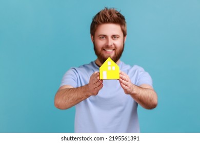 Portrait Of Happy Bearded Man Holding In Hands Little Yellow Paper House Looking At Camera With Pleasant Smile, Dreaming About Own House. Indoor Studio Shot Isolated On Blue Background.