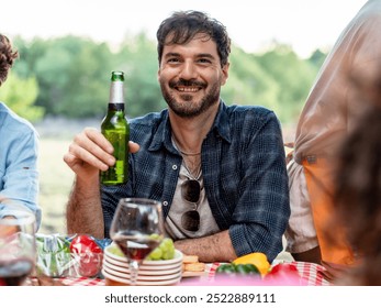 Portrait of a happy bearded man at an outdoor gathering, holding a beer bottle and smiling. Friends enjoying picnic in nature with food and drinks on the table. - Powered by Shutterstock