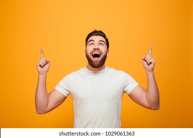 Portrait of a happy bearded man pointing fingers up at copyspace isolated over yellow background