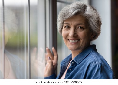 Portrait Of Happy Beautiful Aged 50s Female Of Hispanic Ethnicity Stand By Large Window Look At Camera. Head Shot Of Attractive Healthy Senior Latin Woman Enjoy Active Life In Retirement. Copy Space