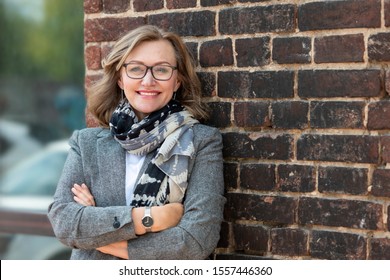 Portrait Of A Happy Beautiful Blonde, 55 Years Old, Smiling. Outdoors, In A City On The Background Of The Old Wall Of Red Brick.