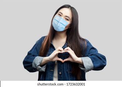 Portrait of happy beautiful brunette asian young woman with surgical medical mask in blue denim jacket standing with love heart gesture with smile. studio shot, indoor isolated on grey background