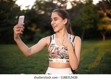 Portrait Of A Happy Beautiful Brunette Sports Fitness Woman In Top With Tropical Print Sitting Outdoors In The Park On Grass With Sundown Light Take A Selfie By Mobile Phone Showing Thumbs Up.