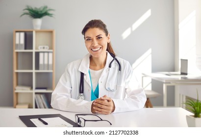 Portrait of happy beautiful Caucasian female doctor who has her own practice. Smiling European physician or cardiologist in white lab coat uniform sitting at desk in her modern office - Powered by Shutterstock