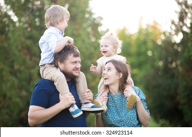 Portrait of happy beautiful family of four walking in park in summer. Mom and dad carrying two little cheerful laughing kids on shoulders. Parents and siblings playing, having fun together - Powered by Shutterstock