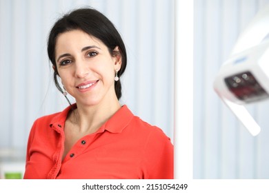 Portrait Of Happy Beautiful Female Doctor, Attractive Pretty Middle Aged Caucasian Armenian Ethnic Dentist In Red Polo Standing In Dental Office At Clinic, Smiling, Looking At Camera. 