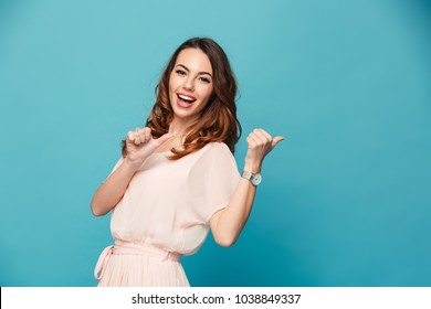 Portrait of a happy beautiful girl wearing dress pointing fingers away at copy space isolated over blue background