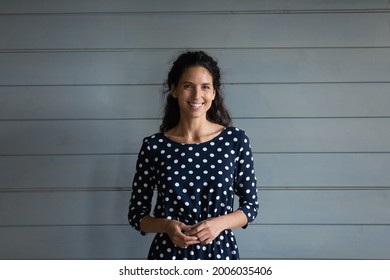 Portrait of happy beautiful Latin millennial 30s woman wearing spotted dress, standing against grey background with toothy smile, looking at camera and laughing. Head shot, profile picture