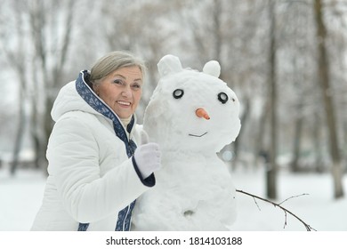 Portrait Of Happy Beautiful Senior Woman Posing In Snowy Winter Park With Snowman