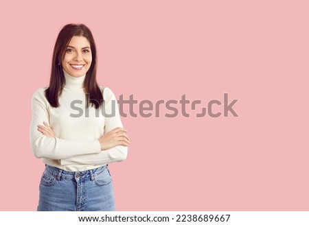 Portrait of happy beautiful smiling charming young brunette woman in casual white turtleneck and blue jeans standing with her arms crossed on empty solid light pink copy space studio background
