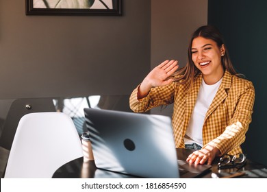 Portrait of happy beautiful stylish brunette young woman sitting at table, looking at laptop screen on video call and greeting with waving. Indoor studio shot, cafe, office background. - Powered by Shutterstock