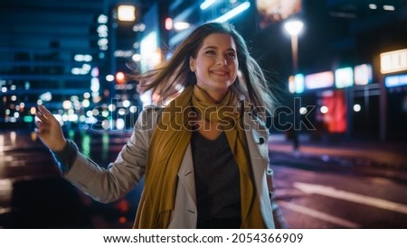Portrait of a Happy Beautiful Woman in Trench Coat Walking in a Modern City Street with Neon Lights at Night. Attractive Female Jumping Around Smiling, Excited to Be in an Urban Cinematic Environment.