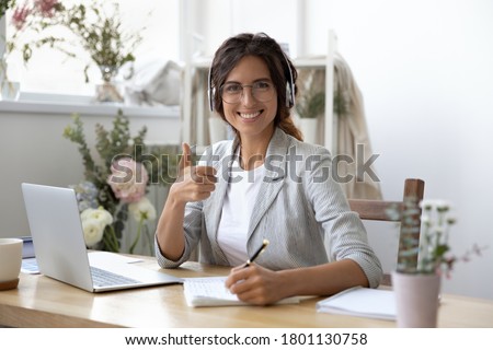 Portrait of happy beautiful young businesswoman in glasses wearing headphones, enjoying listening educational webinar in modern creative workplace, showing thumbs up gesture, recommending seminar.