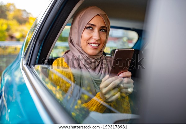 Portrait of happy beautiful young Muslim woman in\
beige hijab sitting on backseat of car. Beautiful Muslim business\
woman is using a smart phone and smiling while sitting on back seat\
in the car