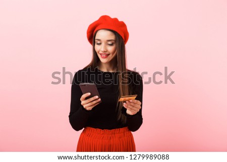 Portrait of a happy beautiful young woman wearing red beret standing isolated over pink background, holding mobile phone and plastic credit card